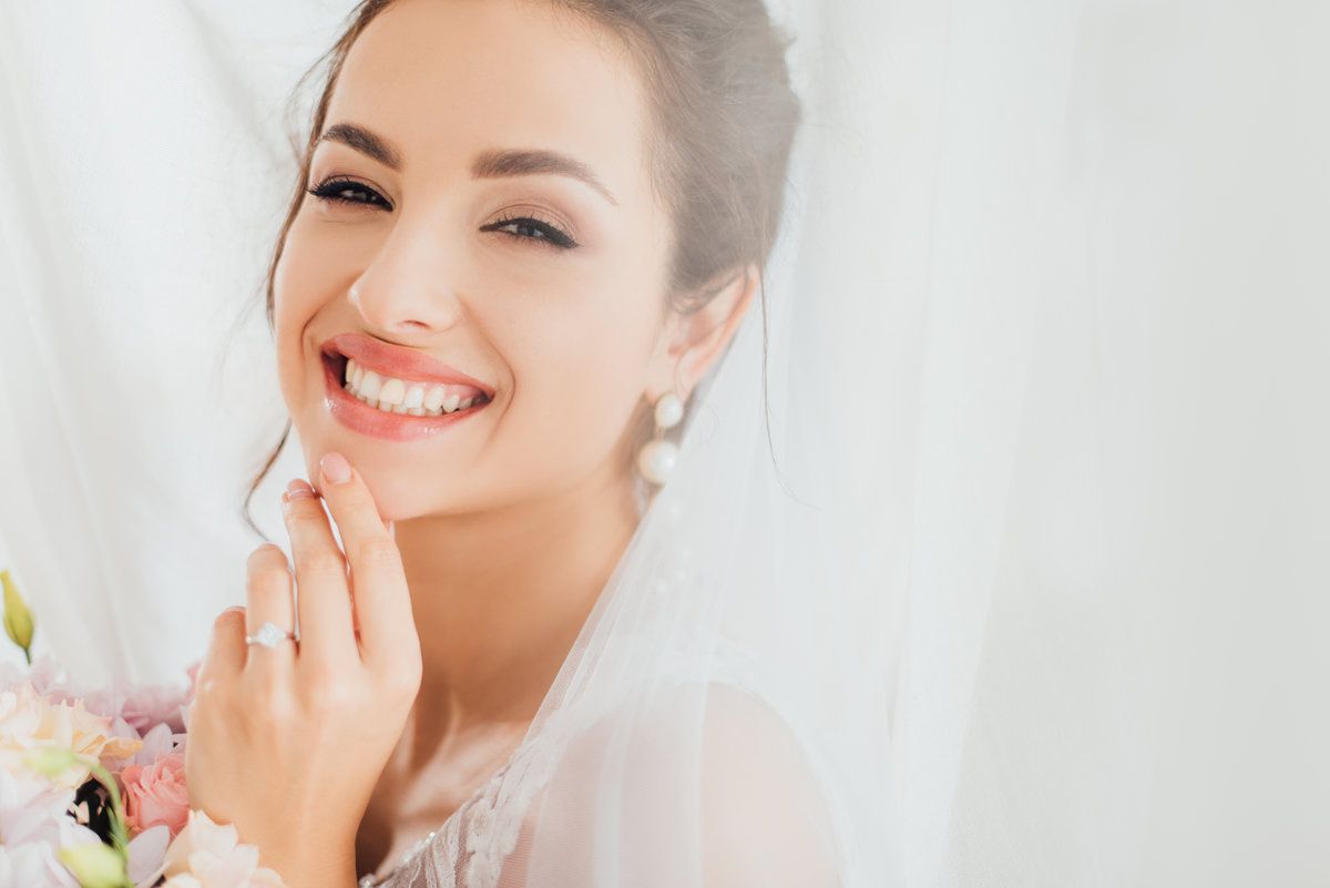 Selective focus of young bride looking at camera near bouquet and curtains